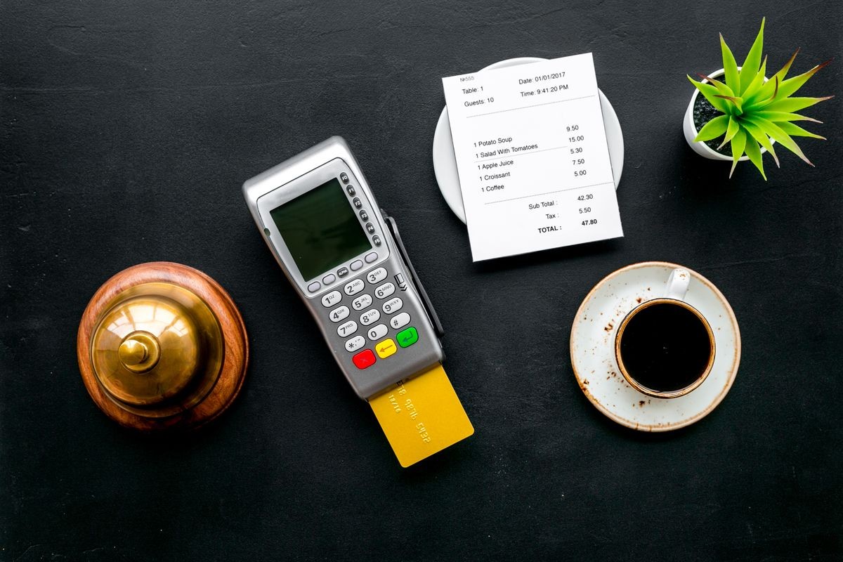 Electronic payments. Pay the bill by card concept. Bank card inserted in payment terminal near bill, service bell, coffee on black background top view